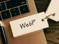 Notatka z napisem "WebP" na karcie przyczepionej do breloczka, leżąca na klawiaturze laptopa obok długopisu.