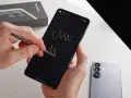 Ręce trzymają smartfon, rysując rysikiem na czarnym ekranie. Obok leży nowy Samsung S26, gotowy na premierę.