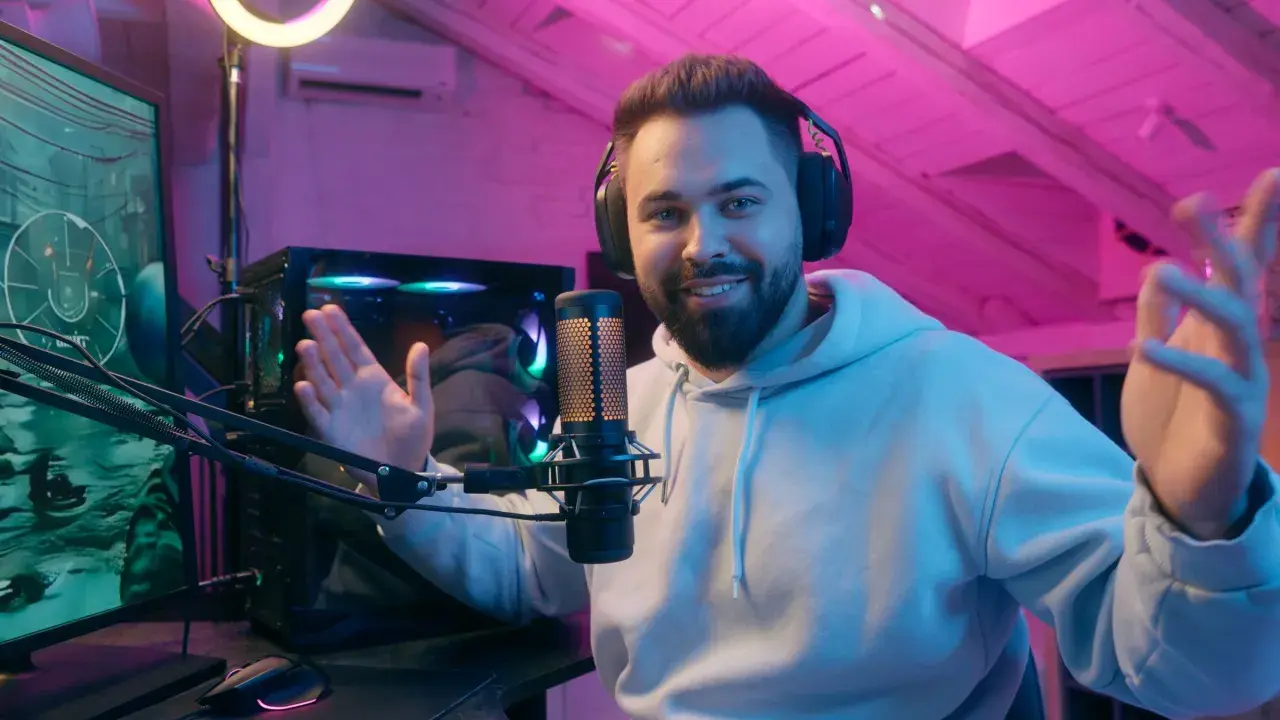 Uśmiechnięty streamer w słuchawkach, przed komputerem i mikrofonem, tłumaczy, streaming co to jest, w pokoju z różowym oświetleniem.