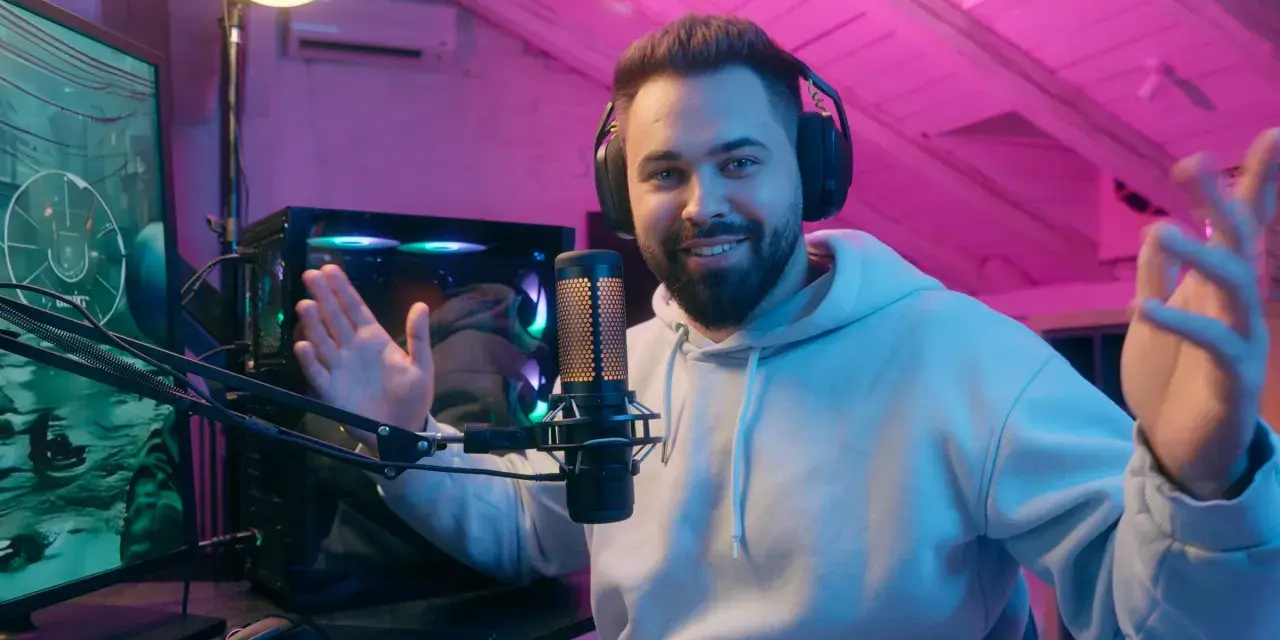 Uśmiechnięty streamer w słuchawkach, przed komputerem i mikrofonem, tłumaczy, streaming co to jest, w pokoju z różowym oświetleniem.