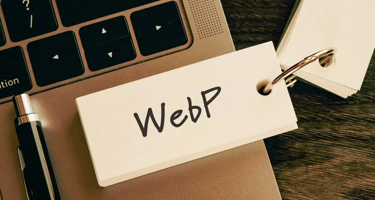 Notatka z napisem "WebP" na karcie przyczepionej do breloczka, leżąca na klawiaturze laptopa obok długopisu.