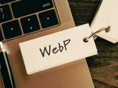 Notatka z napisem "WebP" na karcie przyczepionej do breloczka, leżąca na klawiaturze laptopa obok długopisu.