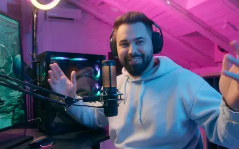 Uśmiechnięty streamer w słuchawkach, przed komputerem i mikrofonem, tłumaczy, streaming co to jest, w pokoju z różowym oświetleniem.