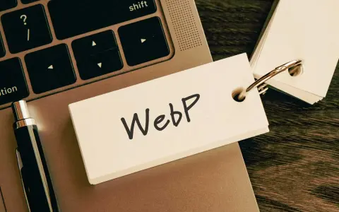 Notatka z napisem "WebP" na karcie przyczepionej do breloczka, leżąca na klawiaturze laptopa obok długopisu.
