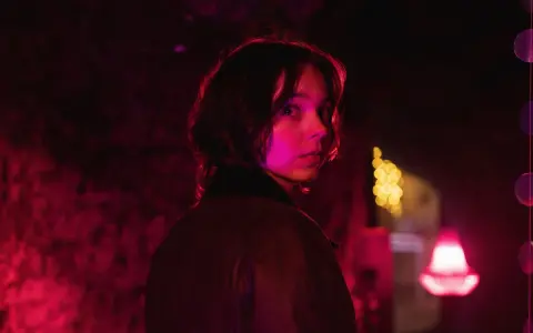 Młoda kobieta z zaciekawieniem spogląda przez ramię w mrocznym, neonowym otoczeniu. Czy to scena z nowego serialu Netflix?