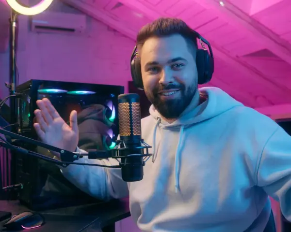 Uśmiechnięty streamer w słuchawkach, przed komputerem i mikrofonem, tłumaczy, streaming co to jest, w pokoju z różowym oświetleniem.