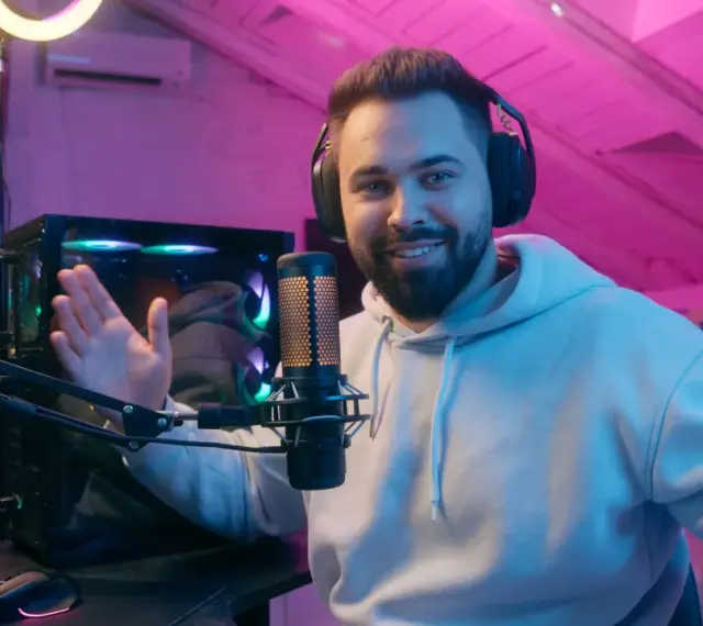 Uśmiechnięty streamer w słuchawkach, przed komputerem i mikrofonem, tłumaczy, streaming co to jest, w pokoju z różowym oświetleniem.