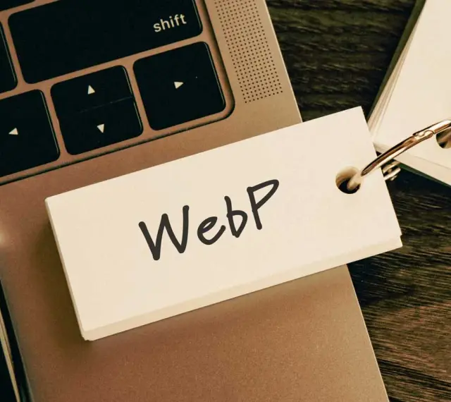 Notatka z napisem "WebP" na karcie przyczepionej do breloczka, leżąca na klawiaturze laptopa obok długopisu.
