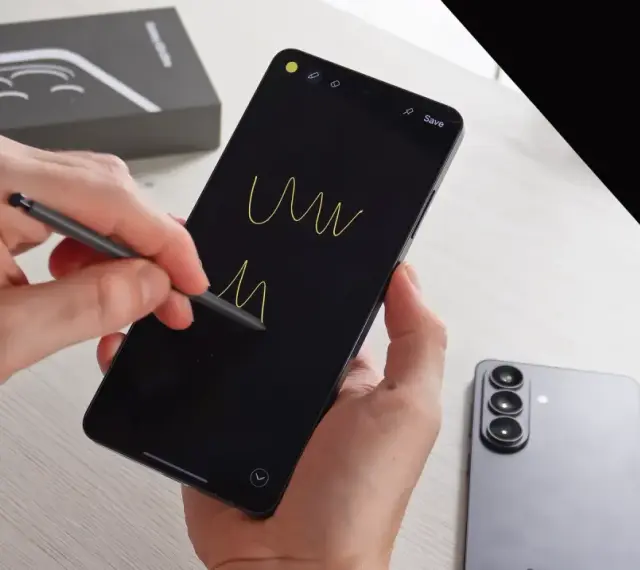 Ręce trzymają smartfon, rysując rysikiem na czarnym ekranie. Obok leży nowy Samsung S26, gotowy na premierę.