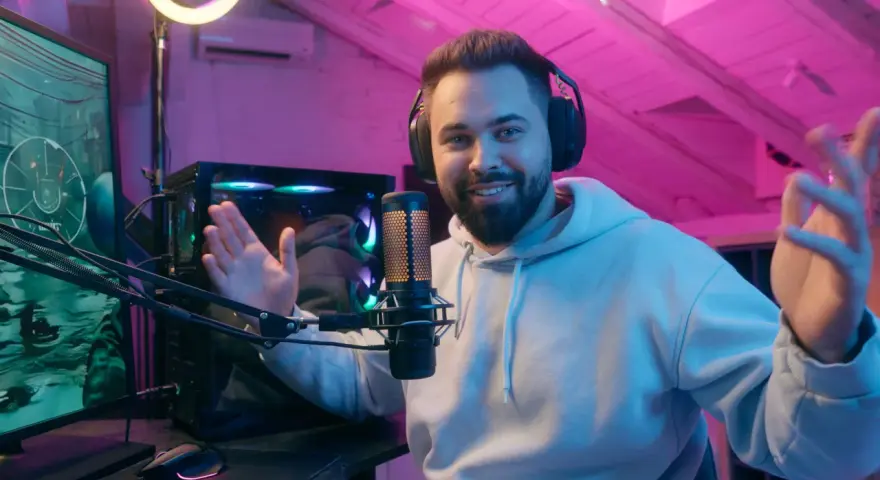Uśmiechnięty streamer w słuchawkach, przed komputerem i mikrofonem, tłumaczy, streaming co to jest, w pokoju z różowym oświetleniem.