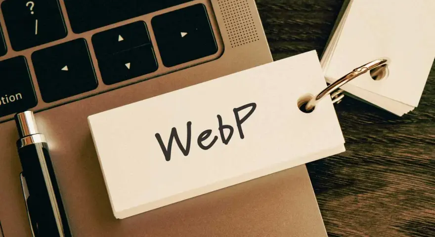 Notatka z napisem "WebP" na karcie przyczepionej do breloczka, leżąca na klawiaturze laptopa obok długopisu.