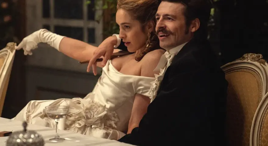 Elegancka para przy stole, zapowiedź nowych produkcji Netflix. Kobieta w białej sukni z rękawiczkami, mężczyzna w czarnym fraku.
