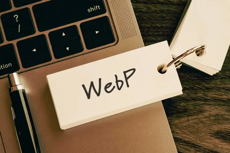 Notatka z napisem "WebP" na karcie przyczepionej do breloczka, leżąca na klawiaturze laptopa obok długopisu.
